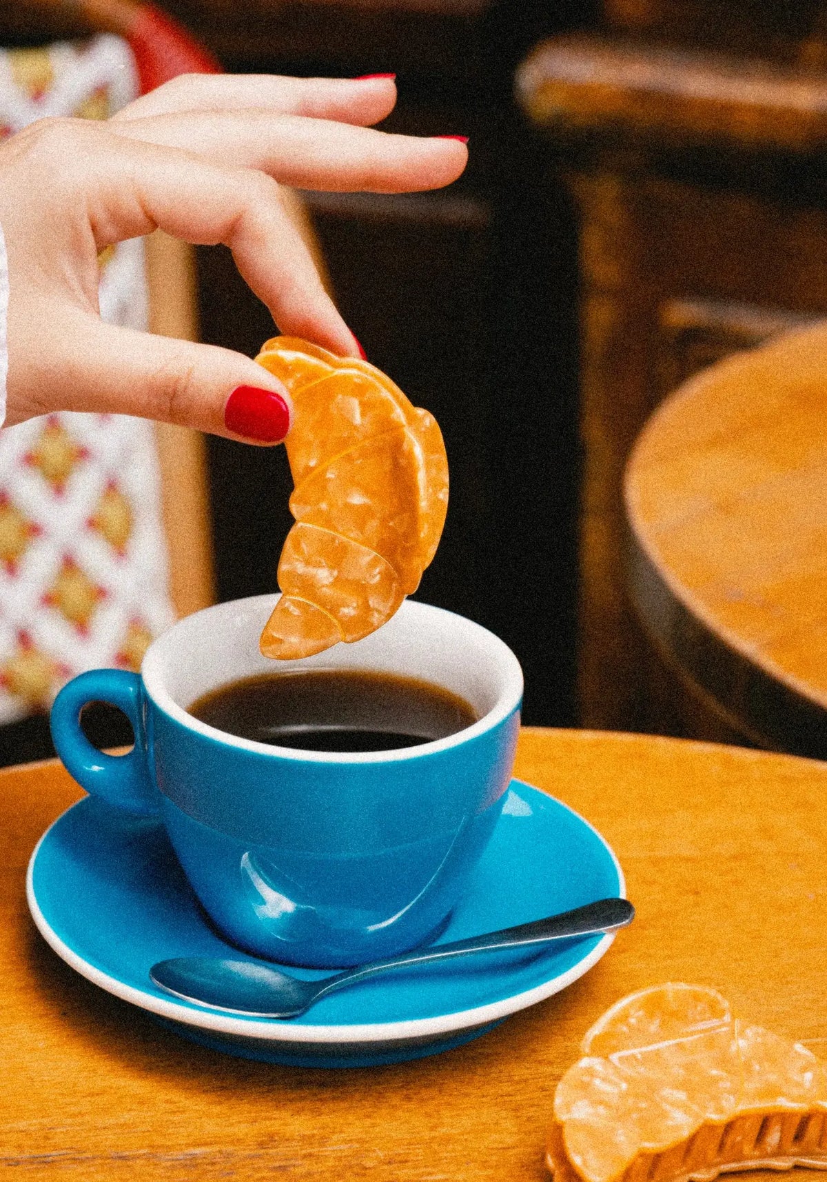 Hand som doppar en croissantformad hårklämma från Coucou Suzette i en blå kaffekopp på ett kafébord
