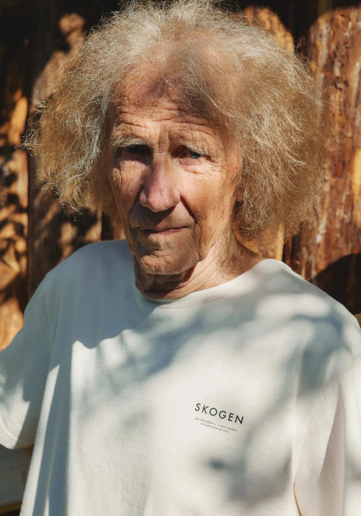 Jan Håfström med yvigt ljust hår i benvit t-shirt med texten SKOGEN på bröstet, framför en solbelyst trävägg.