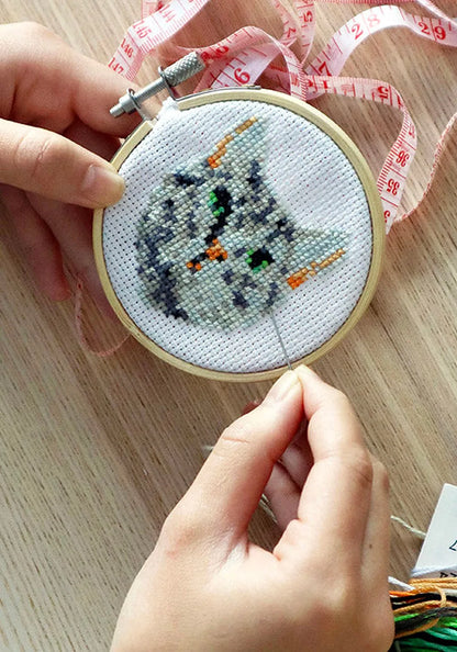Händer som broderar en grårandig katt i korsstygn i en liten broderiram i bambu, på ett träbord med måttband och broderigarn.