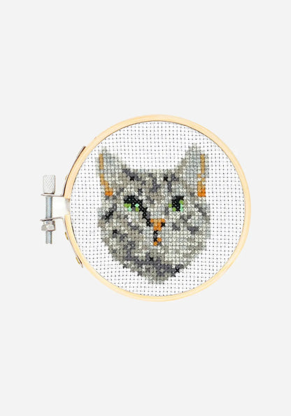Färdigt korsstygnsbroderi i en broderiram i bambu, med ett grårandigt kattansikte med gröna ögon och orange nos.