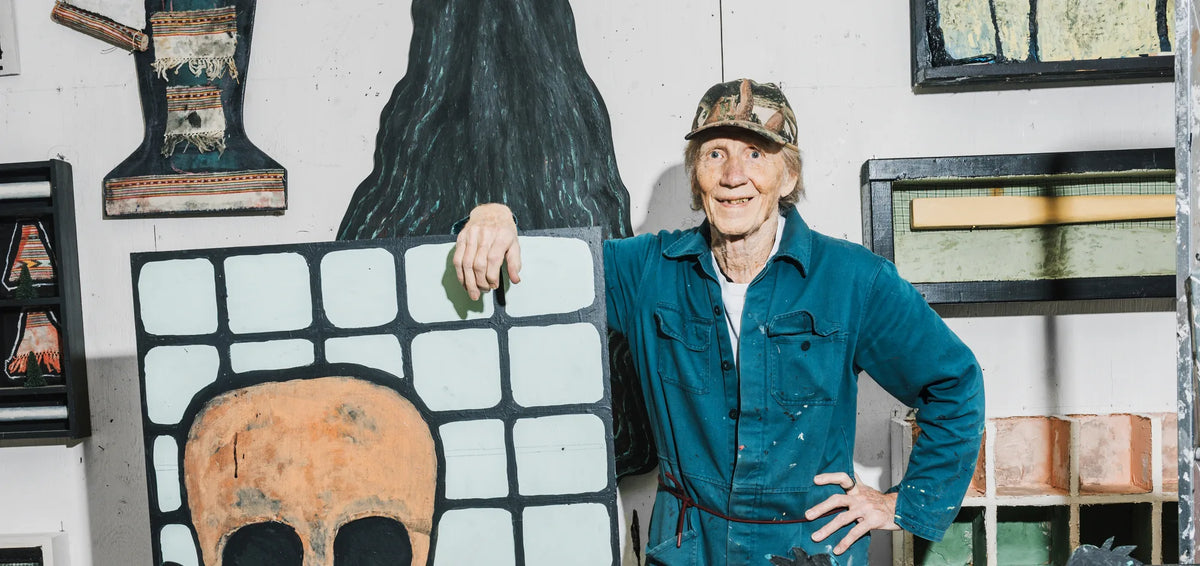 Jan Håfström i blå overall och keps i sin ateljé, lutad mot en målning med en dödskalle bakom ett svart rutnät, med en installation av svarta palmer i förgrunden.