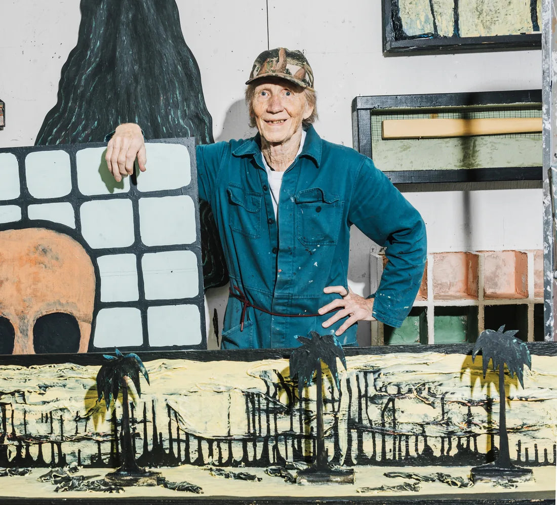 Jan Håfström i blå overall och keps i sin ateljé, lutad mot en målning med en dödskalle bakom ett svart rutnät, med en installation av svarta palmer i förgrunden.