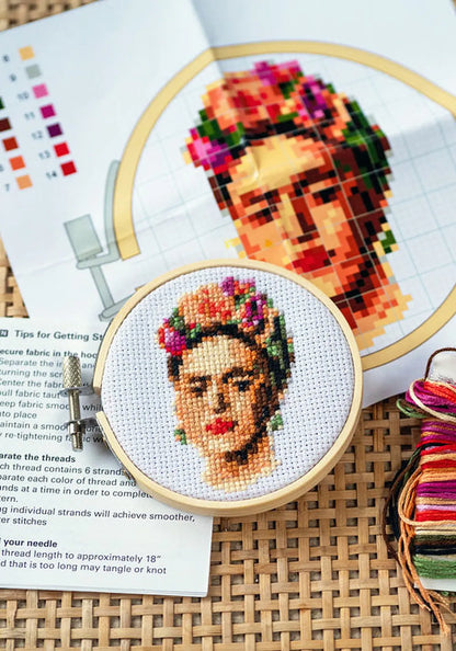 Innehållet i ett broderikit med Frida Kahlo: mönster, instruktioner, en broderiram med färdigt broderi och broderigarn i många färger, på en flätad yta.