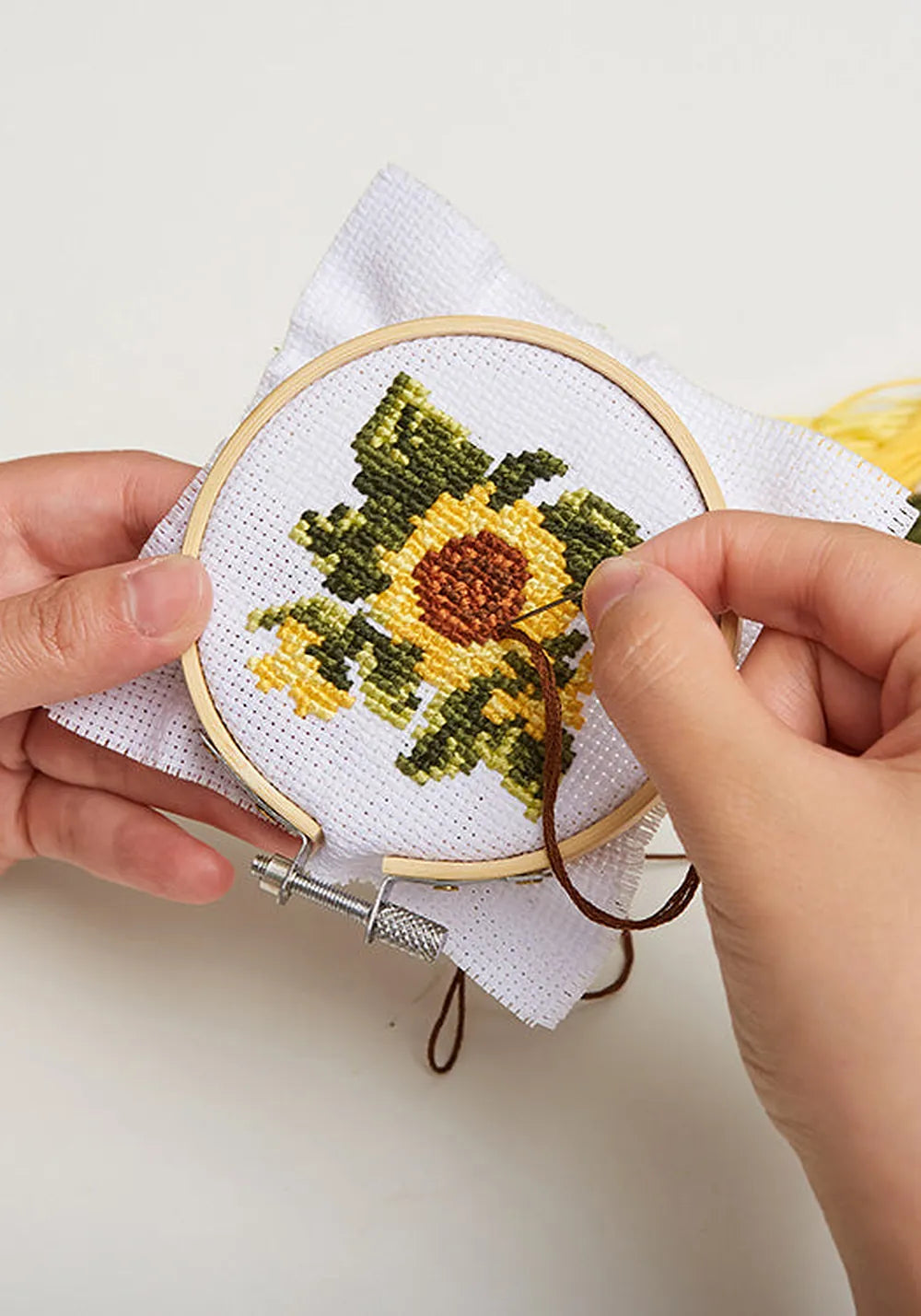 Händer som broderar en gul solros i korsstygn i en liten broderiram i bambu, med garn i bakgrunden.