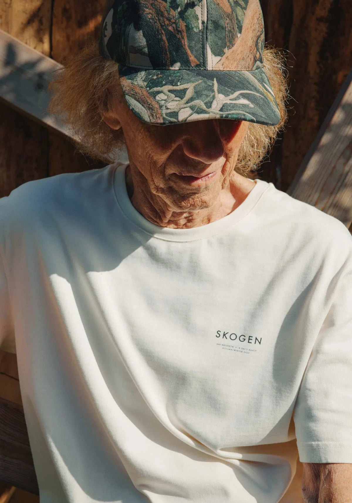 Jan Håfström i benvit t-shirt med texten SKOGEN på bröstet och en keps med skogsmönster, i solljus framför en trävägg.