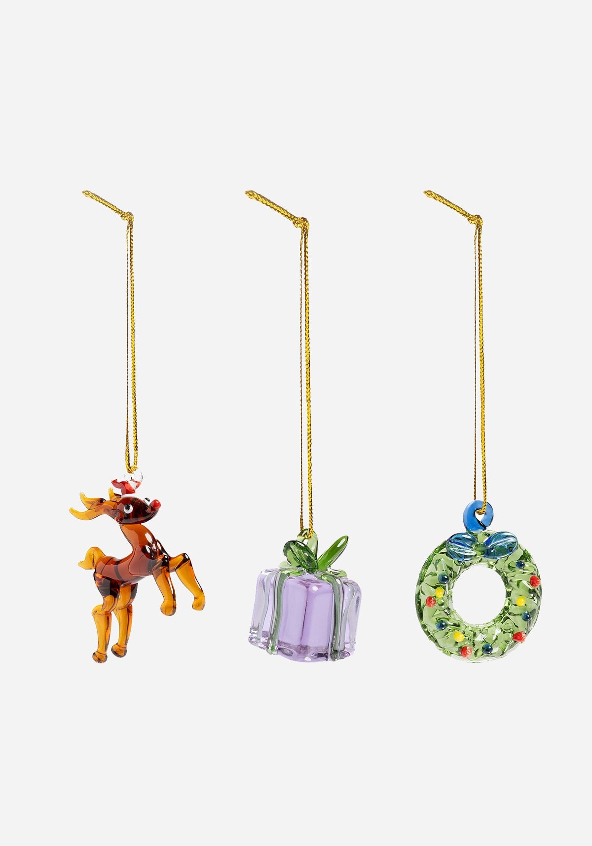 Tre julgranshängen i glas i guldfärgade snören: en brun ren med röd luva, ett lila paket med grön rosett och en grön krans med färgglada prickar.