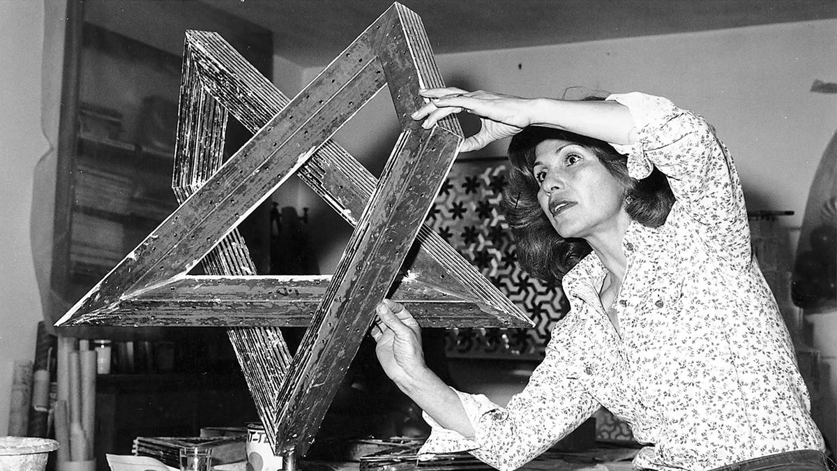 Svartvitt fotografi av Monir Shahroudy Farmanfarmaian i sin ateljé i Teheran, 1975. Hon håller och undersöker en stor geometrisk skulptur uppbyggd av sammanfogade trianglar. I bakgrunden syns ett mönstrat galler och verktyg på en arbetsbänk.