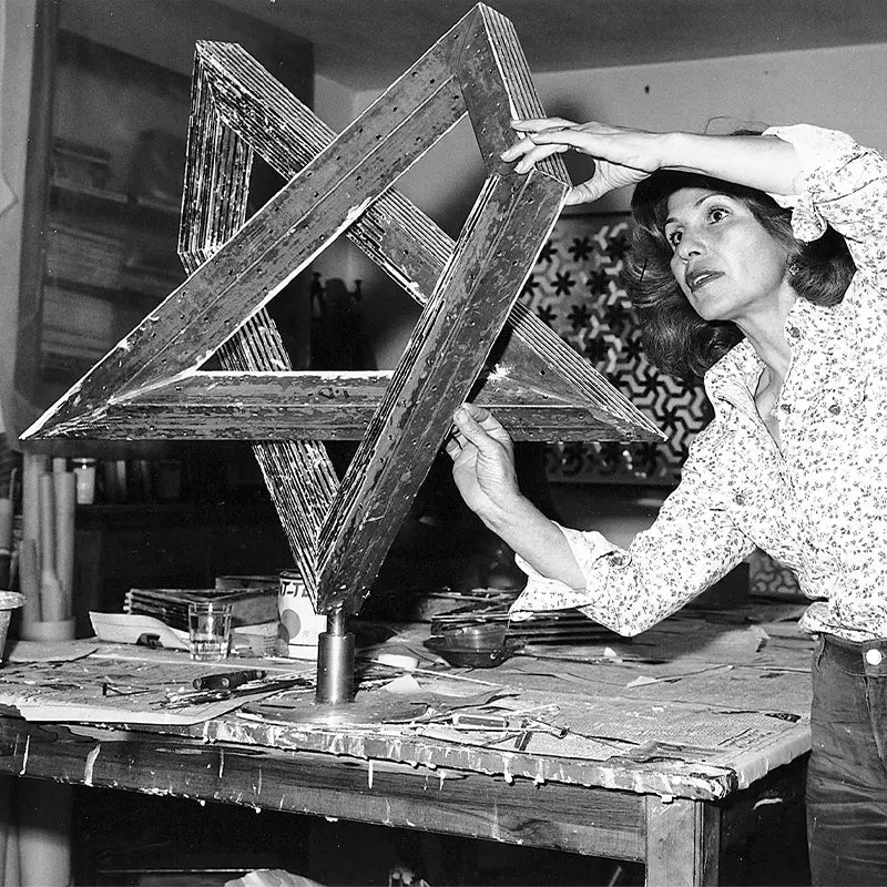 Svartvitt fotografi av Monir Shahroudy Farmanfarmaian i sin ateljé i Teheran, 1975. Hon håller och undersöker en stor geometrisk skulptur uppbyggd av sammanfogade trianglar. I bakgrunden syns ett mönstrat galler och verktyg på en arbetsbänk.
